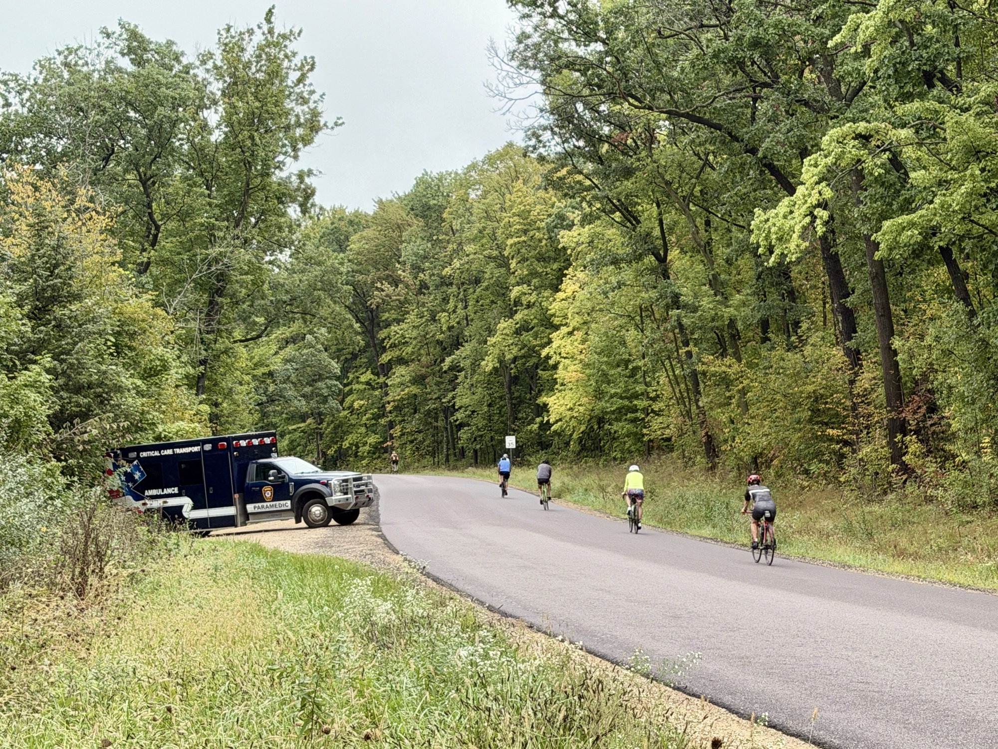 An ambulance providing standby coverage while cyclists travel through a wooded roadway.