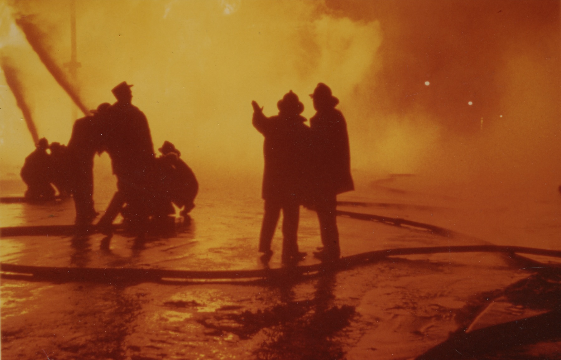 Firefighters silhouetted against the glow of a large nighttime fire.